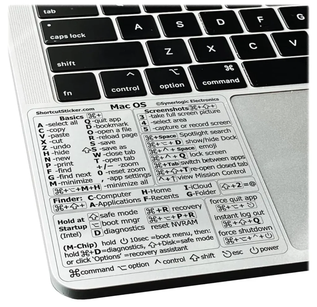 Primer plano de un teclado Mac con una pegatina que muestra atajos de teclado Mac, cubriendo la zona del reposamanos. La pegatina destaca comandos para las funciones básicas, de gestión de ventanas, del Finder, de inicio y del sistema.