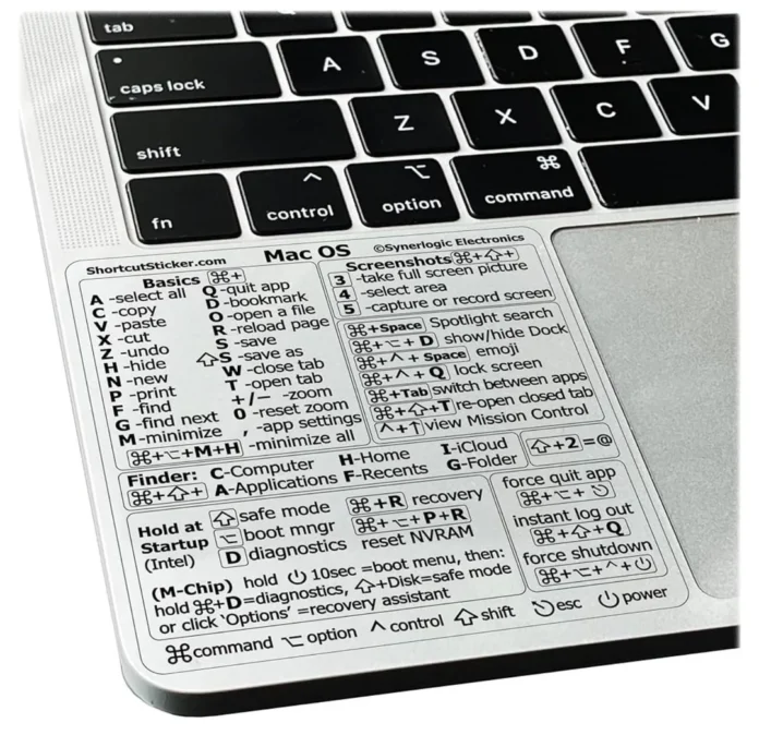 Primer plano de un teclado Mac con una pegatina que muestra atajos de teclado Mac, cubriendo la zona del reposamanos. La pegatina destaca comandos para las funciones básicas, de gestión de ventanas, del Finder, de inicio y del sistema.