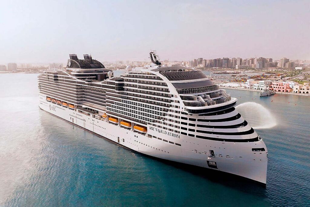 Un crucero grande y moderno navega cerca de una ciudad costera, con varias cubiertas y botes salvavidas a la vista. Las aguas tranquilas reflejan el perfil de los edificios del fondo bajo un cielo despejado: una escena imprescindible para cualquier viajero.