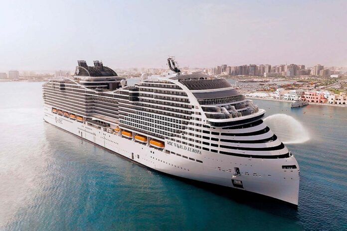 Un crucero grande y moderno navega cerca de una ciudad costera, con varias cubiertas y botes salvavidas a la vista. Las aguas tranquilas reflejan el perfil de los edificios del fondo bajo un cielo despejado: una escena imprescindible para cualquier viajero.