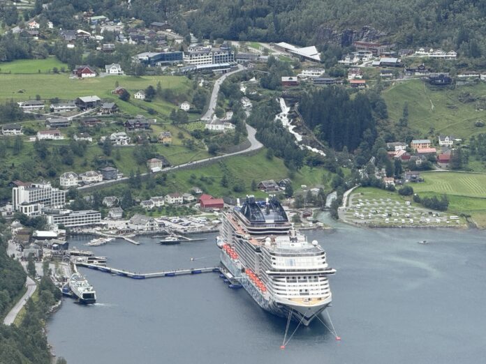 Un gran crucero, el MSC Euribia, está atracado en el puerto de un pintoresco pueblo costero con verdes colinas, casas dispersas, carreteras serpenteantes y una cascada al fondo: un lugar idílico para cualquier aficionado a los clubes náuticos.
