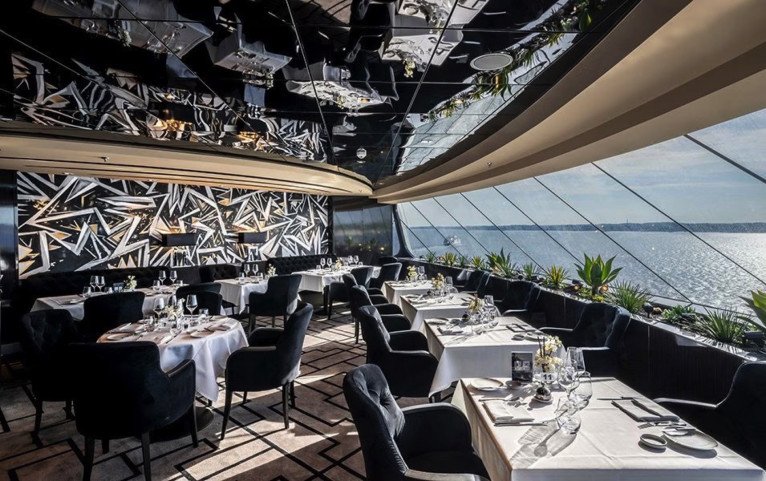Elegante restaurante con sillas negras y manteles blancos, grandes ventanales que ofrecen una vista panorámica del agua, arte geométrico en las paredes y un techo de espejos que refleja la luz natural, perfecto para los huéspedes del Yacht Club a bordo del crucero MSC Euribia.